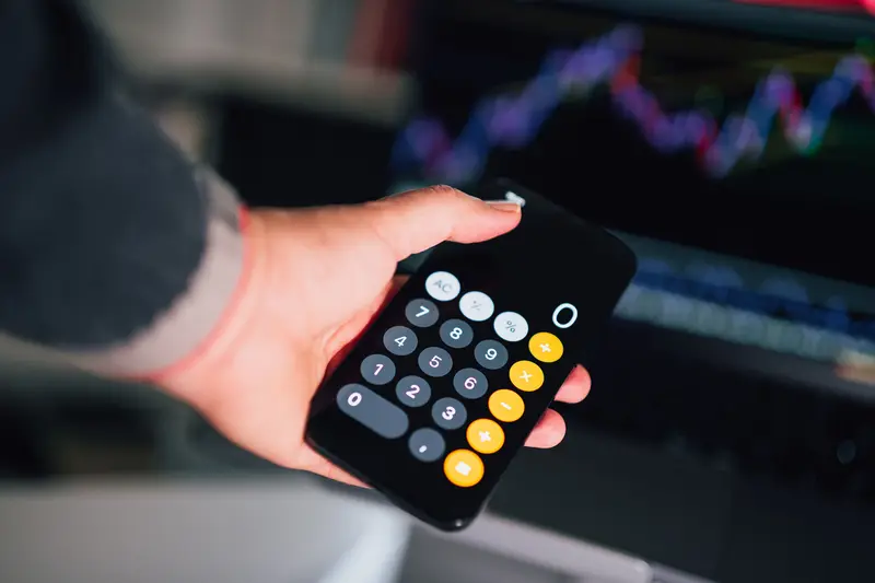 Hand holding a phone calculator in front of a glowing stock chart